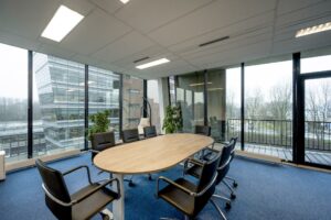 Vergaderruimte met ovale houten tafel, zwarte stoelen en uitzicht op kantoren aan de Joop Geesinkweg.